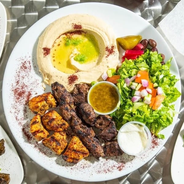 Grilled chicken and meat skewers with hummus, fresh salad, pickles, and dipping sauces on a white plate.