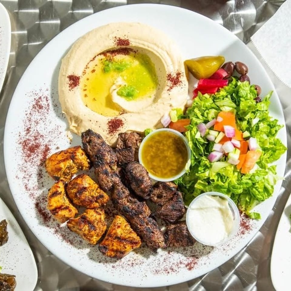 Grilled chicken and meat skewers with hummus, fresh salad, pickles, and dipping sauces on a white plate.