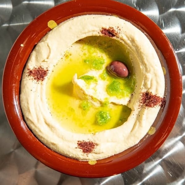 Creamy hummus in a red bowl, topped with olive oil, green sauce, an olive, and red spice.