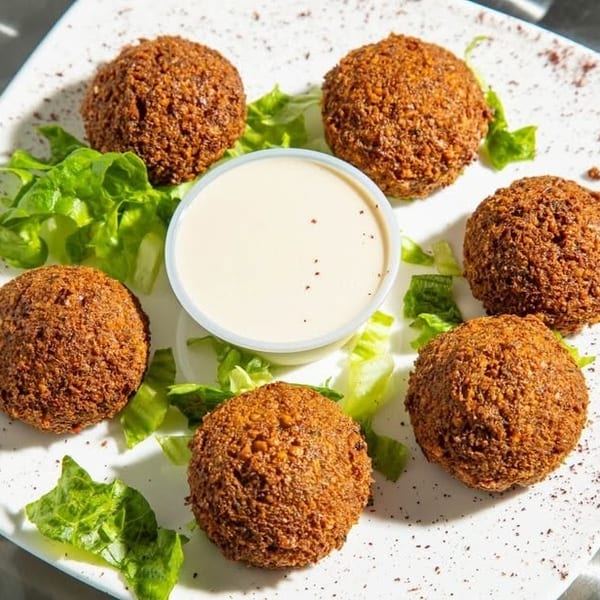Six crispy falafel balls with a side of tahini sauce and lettuce on a white plate.