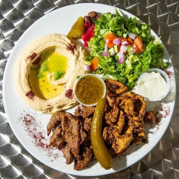 A large plate of Middle Eastern food with hummus, salad, beef and chicken shawarma, pickles, and sauces.
