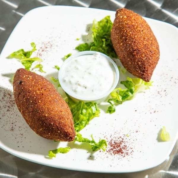 Two golden-brown fried kibbeh served with a creamy white dipping sauce and fresh lettuce.